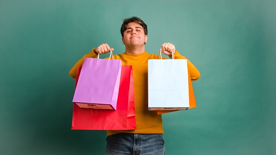 Smiling young Hispanic male looking at camera while standing with toothy smile showing shopping bags in both moving up and down hands against green wall