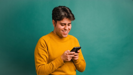 Smiling Hispanic young man in yellow outfit browsing smartphone while standing against green background
