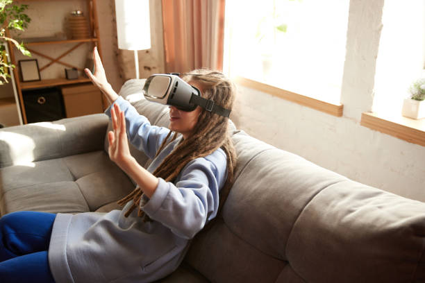 istock Excited young lady with long dreadlocks playing virtual reality game in glasses sitting on sofa in living room at home. 2010560389