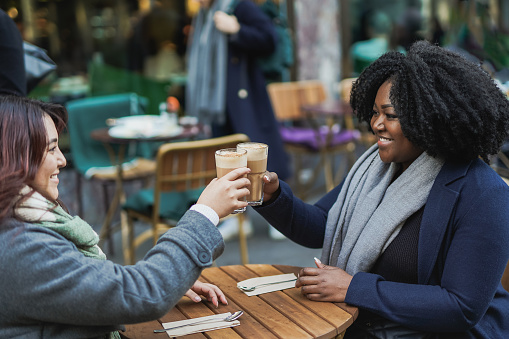 Happy african and asian girl cheering together with coffee outside of vintage bar - Multiracial female friends having fun meeting in the city during winter time