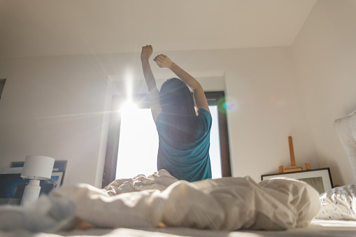 Young woman waking up in the morning, her arms are raised, she is stretching, sunbeam from the window is falling on her while she is sitting on her bed in bedroom