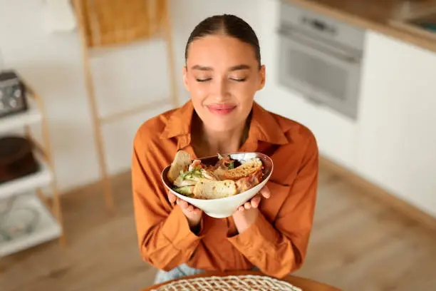 Contented woman in blouse cherishes the aroma of her fresh salad bowl, sitting in minimalist kitchen with wooden floor and modern appliances