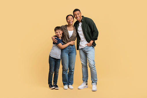 Portrait of happy cute african american family father, mother and teenager boy son posing isolated on beige studio background, embracing, smiling at camera, full length shot, copy space