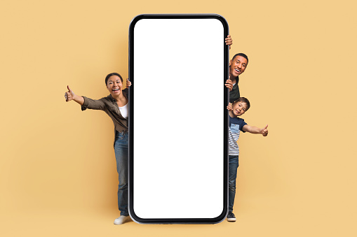 Entertaining mobile application. Excited young black family parents and son standing behind big smartphone with white screen, beige background. Cellphone display mockup, free copy space