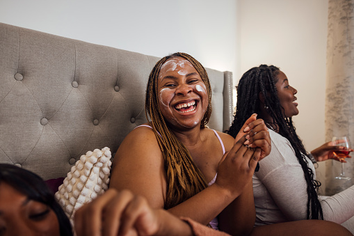 A group of female friends wearing pyjamas, spending the self-care night together in one of their flats in Newcastle upon Tyne, England. They are all sitting on a bed after doing their skincare while laughing, the main focus is a woman looking at the camera and smiling with a cream on her face.
