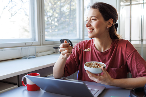 Photo of a young woman eating a healthy breakfast and looking through the window while using a laptop