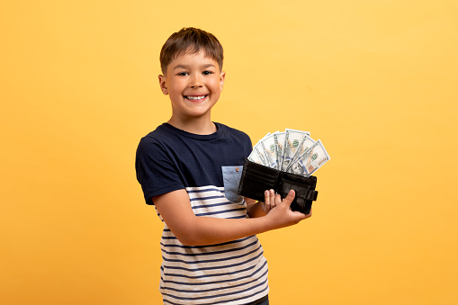 Pocket money, kids savings, financial literacy for children. Cheerful cute preteen kid handsome boy in casual outfit showing leather wallet full of cash dollar banknotes, smiling, yellow background