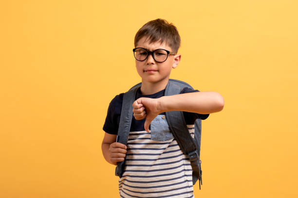 Unhappy handsome preteen boy schooler showing thumb down Unhappy handsome preteen boy schooler wearing eyeglasses with backpack showing thumb down, isolated on yellow background. Sad child doesnt like his new school, copy space thumb down stock pictures, royalty-free photos & images