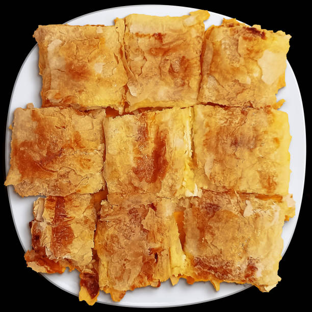 Bunch of freshly oven baked Greek Cheese Pie square pieces, prepared with Feta and Curd Cheese, served in white ceramic plate, isolated on black background.