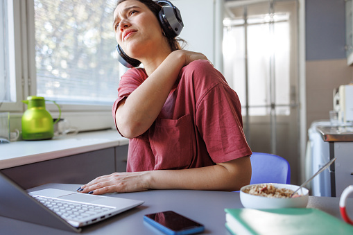 A young woman suffering from neck pain while working on the laptop at home. She is self-massaging her neck and shoulders.