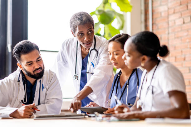 Medical Training A female physician checks in on some medical students who are seated together at a table working on a project. They are each dressed professionally and are focused on the task as they work together. medical occupation stock pictures, royalty-free photos & images