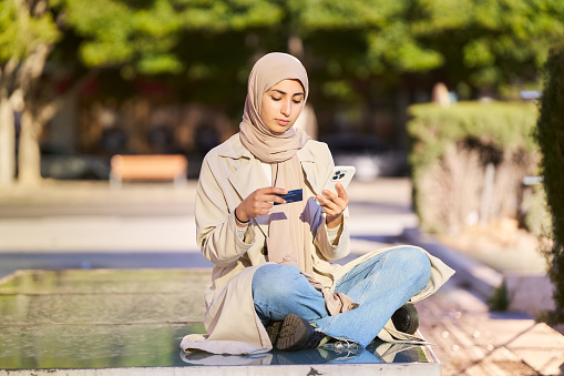 arab woman in headscarf pays for online purchases with her credit card via mobile phone