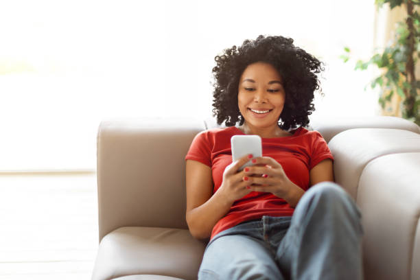istock Cheerful young black woman sitting on couch and using her smartphone 2010781115