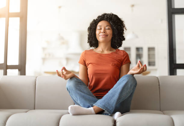 Stress Relief Concept. Peaceful young black woman sitting cross-legged on couch Stress Relief Concept. Peaceful young black woman in casual clothes sitting cross-legged on couch, smiling calm african american female practicing meditation with closed eyes, enjoying home comfort managing stress stock pictures, royalty-free photos & images