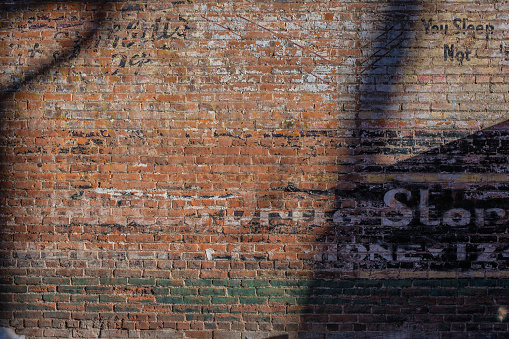 A partly shaded, weathered red brick wall with old faded writing on it.