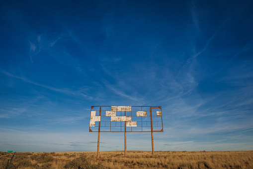 A centered rusty frame of a billboard with a few pieces still attached, in a dry grassland under blue sky.