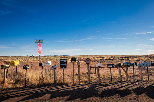 Funky, mismatched mailboxes side by side along the edge of a rural road with a stop sign, against dry grass and blue sky.