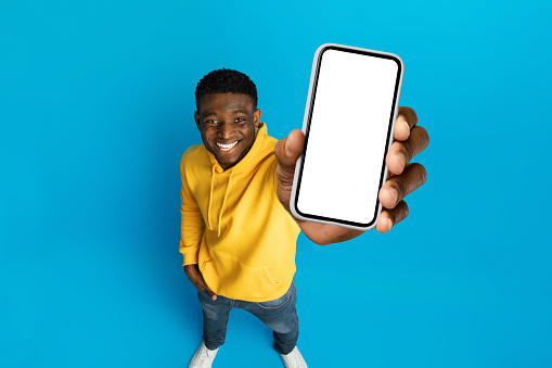 Great offer, newest mobile app, exciting online deal. Cool young african american guy showing phone with white blank mockup screen, isolated on blue studio background, high angle view