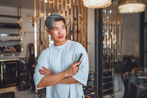 Close up of asian professional Hairdresser or hairstylist man standing confidence with arm crossed with smile and holding hairdressing equipment while looking at camera in beauty salon and barber shop