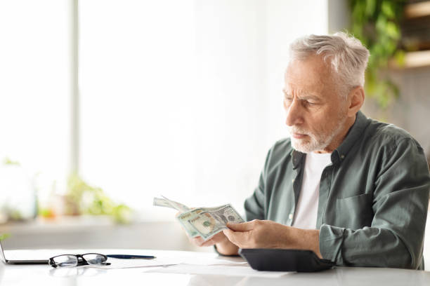 istock Senior male counting dollar cash while sitting at table in kitchen 2010826459