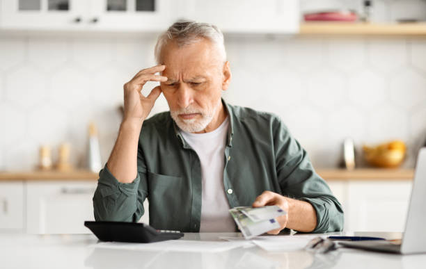 Anxious senior man managing finances, holding cash and using calculator at home