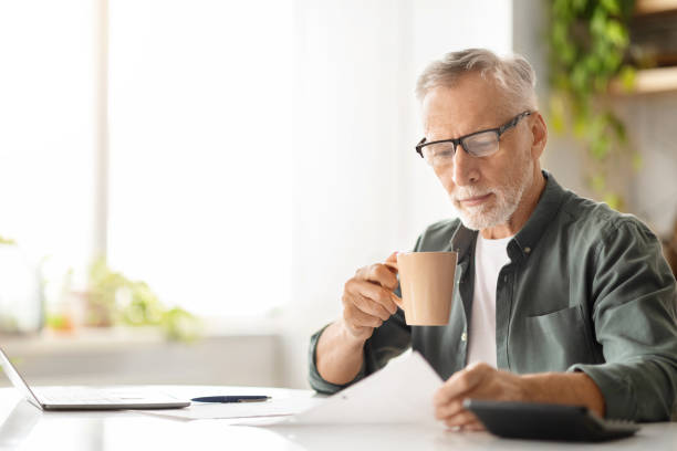 istock Concentrated elderly male in glasses drinking coffee while reviewing documents at home 2010826965