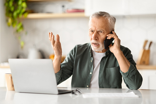 Stressed Senior Man Talking On Cellphone And Using Laptop In Kitchen, Anxious Elderly Gentleman Speaking With Customer Support, Having Problems With Computer Or Internet, Closeup Shot