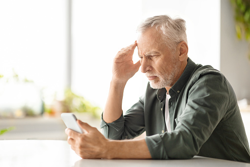 Distressed elderly man suffering headache while looking at smartphone at home, worried senior gentleman reading troubling news or navigating complex app, sitting at table in kitchen, copy space