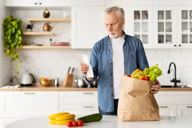 Senior gentleman standing in kitchen, holding paper bag full of groceries while examining shopping receipt, elderly man checking bill, having look of contemplation on face regarding his purchase