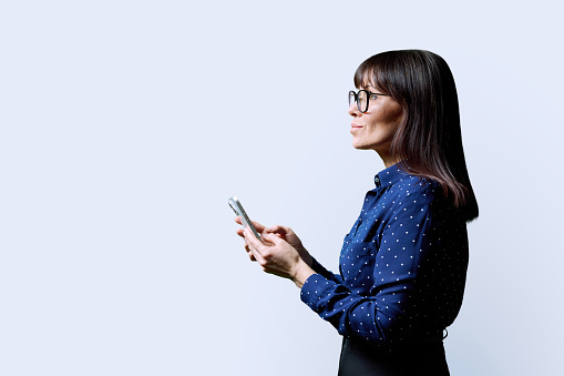 Profile view middle aged smiling woman using smartphone on white background. Mature female looking at phone in hands. Technologies mobile apps applications internet work business leisure communication