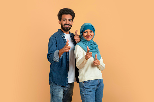 Gotcha. Cheerful Muslim Couple Pointing Fingers At Camera, Indicating Somebody, Happy Arab Man And Woman In Hijab Having Fun Together, Standing Over Beige Studio Background, Copy Space