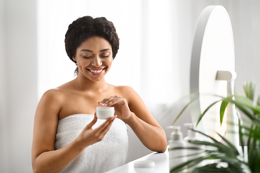 Skin Nourishing. Beautiful Smiling Black Woman Opening Jar With Face Cream, Attractive African American Lady Wrapped In Towel Using Nourishing Cosmetics After Bath, Sitting At Dressing Table At Home