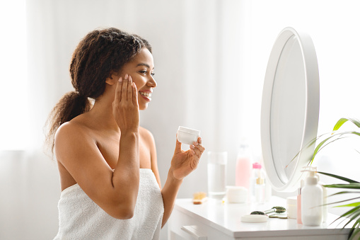 Attractive Young African American Woman Applying Nourishing Cream On Face, Beautiful Black Female Wrapped In Towel Looking In Mirror At Home And Holding Jar With Anti-Aging Cosmetics, Copy Space