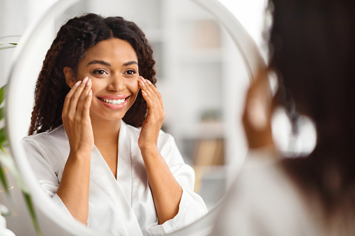 Beautiful Black Young Woman Looking In Mirror And Applying Under Eye Cream, Smiling Attractive African American Woman Wearing White Silk Robe Making Beauty Treatments At Home, Selective Focus