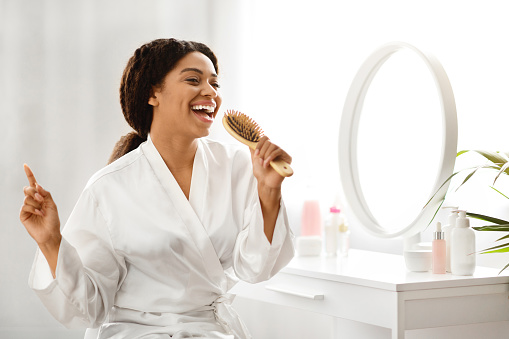 Cheerful African American Woman Singing With Hairbrush While Sitting At Dressing Table In Bedroom, Happy Black Female Having Fun While Brushing Her Hair At Home, Enjoying Haircare Routine