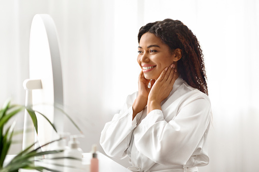 Self-Esteem. Attractive Young Black Woman Looking In Mirror At Home And Touching Face, Happy Beautiful African American Lady Smiling To Her Reflection, Enjoying Appearance, Copy Space