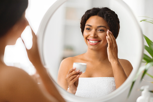 Attractive Black Female Applying Under Eye Cream While Standing Near Mirror At Home, Happy Smiling African American Woman Making Skincare Treatments, Enjoying Daily Beauty Routine, Closeup
