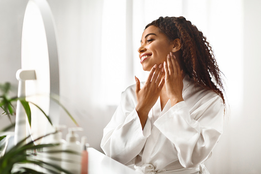 Beautiful African American Woman Looking In Mirror At Her Soft Smooth Skin, Attractive Young Black Female Sitting At Dressing Table And Touching Face, Enjoying Home Skincare Routine, Free Space