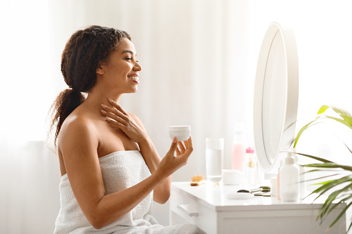 Attractive black woman applying moisturizing cream on her neck while making beauty routine at home, beautiful african american female sitting at dressing table and holding jar with nourishing lotion