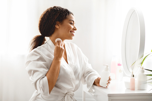 Beautiful Black Female With Cotton Pads Cleansing Neck Skin At Home, Attractive Young African American Woman Sitting At Dressing Table And Looking In Mirror, Making Daily Beauty Routine