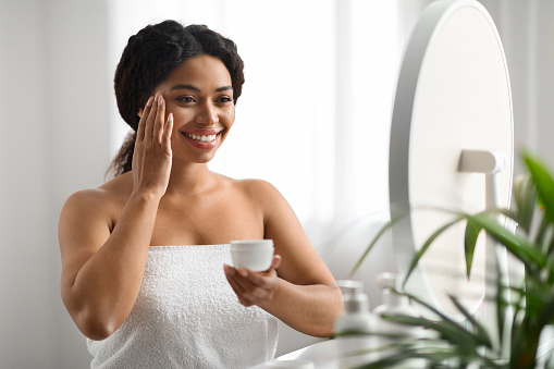 Beautiful Young Black Woman Applying Under Eye Cream At Home, Happy Smiling African American Woman Looking In Mirror While Making Skincare Treatments, Enjoying Daily Beauty Routine, Closeup
