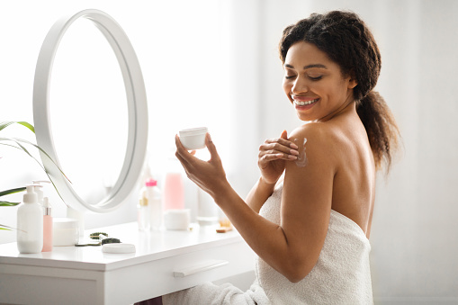 Body Care. Portrait Of Young Black Woman Applying Moisturizing Cream On Shoulder, Beautiful African American Female Using Nourishing Lotion While Making Daily Beauty Routine, Enjoying Skincare