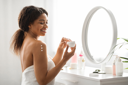 Skin Nourishing. Attractive Black Woman Applying Moisturising Cream Or Lotion On Shoulder, Smiling Beautiful African American Lady Wrapped In Towel After Bath Making Beauty Treatments At Home