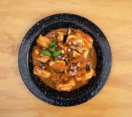 Pasta. Italian food. Top view of traditional stuffed agnolottis with a tomato sauce with mushrooms, bacon and basil in a black dish on the wooden table.