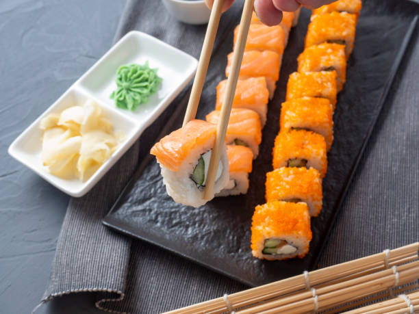 Philadelphia and California rolls on a textured dark plate. One roll is held in Granny sticks above the others. Next to the wasabi and ginger. Side view. Japanese cuisine
