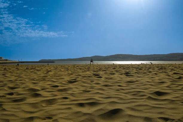 View of sand surface against blue sky background, with clouds, sun halo, reflection on ocean water. Unique natural areas, beaches, deserts. Place where all elements of Nature interact with each other.