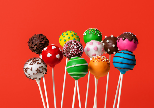 Homemade cake pops with various icings colors, minimalist on a red background.