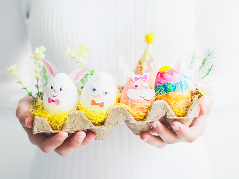 Caucasian girl in a white turtleneck holds in her hands a cardboard box with homemade Easter bunny eggs in yellow straw nests with spring flowers on a white wall background, close-up side view. The concept of crafts, needlework, children art, diy, at home, children creative, artisanal, easter preparation.
