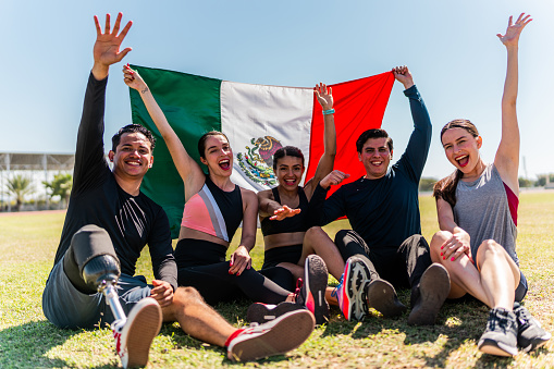 Portrait of athletes holding a mexican flag and celebrating sitting on grass outdoors - Including athletes with disabilities