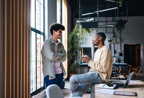 Two young male professionals are laughing and chatting during a break in a bright contemporary workspace.